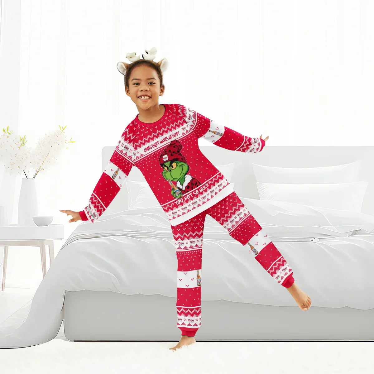 Smiling child in red Coors Light Grinch Christmas family pajamas and reindeer headband in bright bedroom