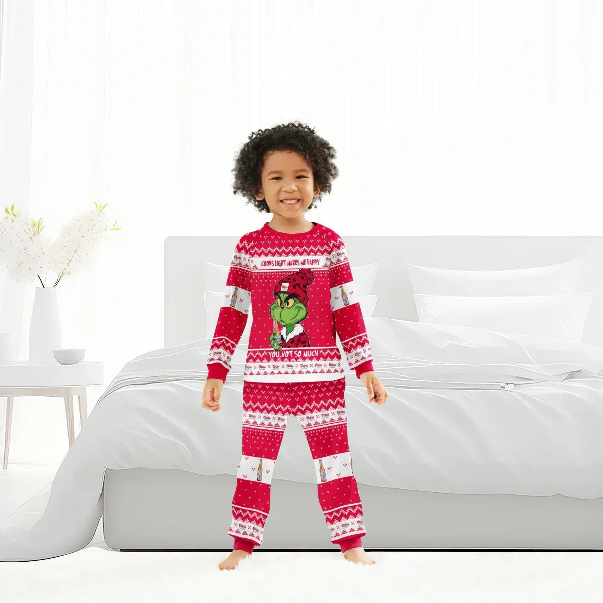 Smiling child in red Coors Light Grinch-themed family pajamas standing barefoot in bright bedroom