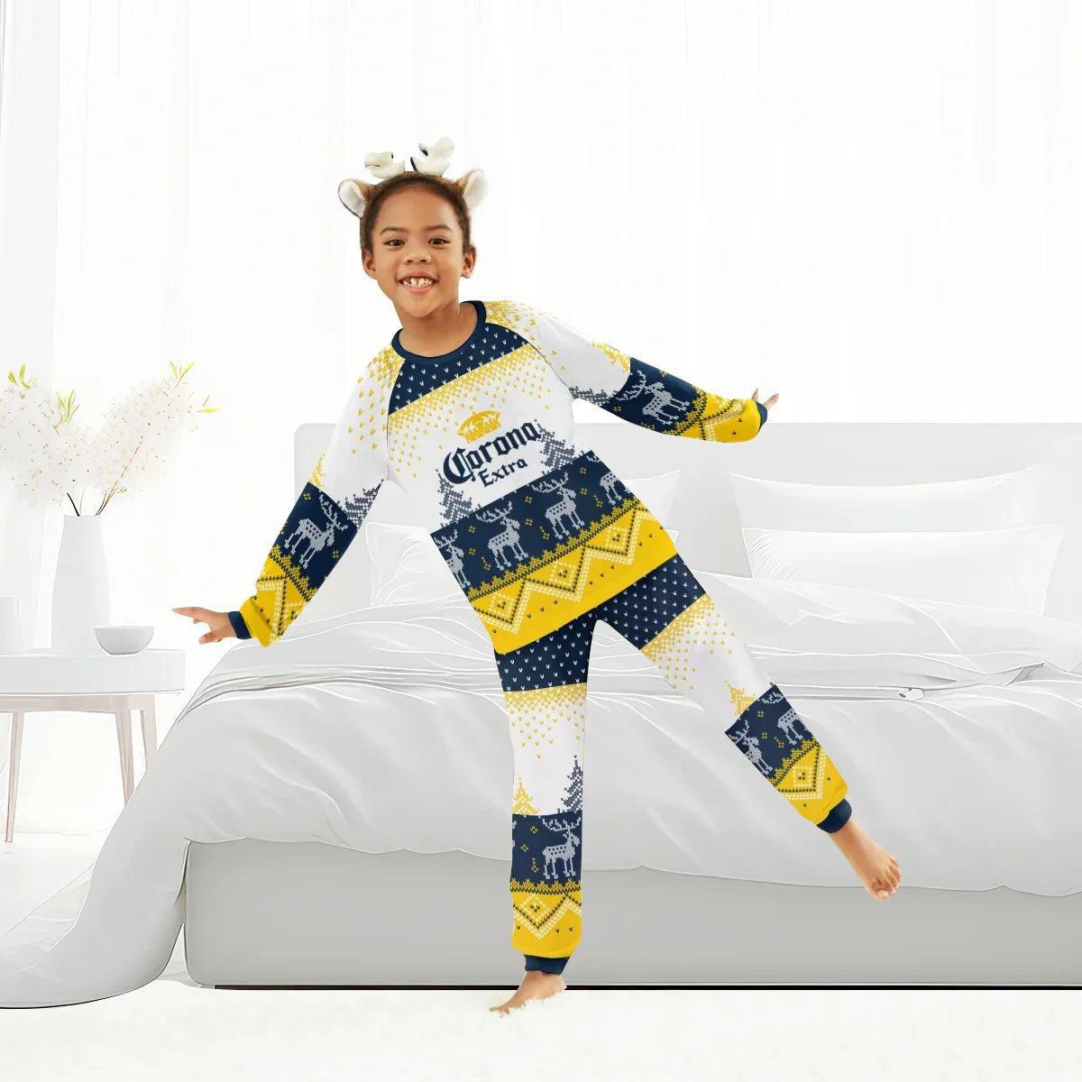Child in Corona Extra reindeer family winter pajamas set and antler headband on bed in bright room