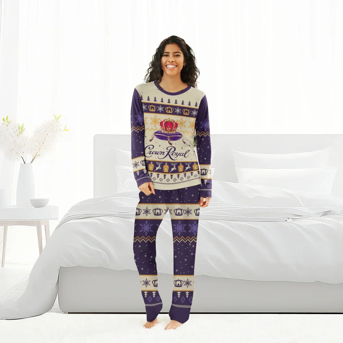 Smiling woman in purple and beige Crown Royal Christmas pajamas standing barefoot in a white bedroom.