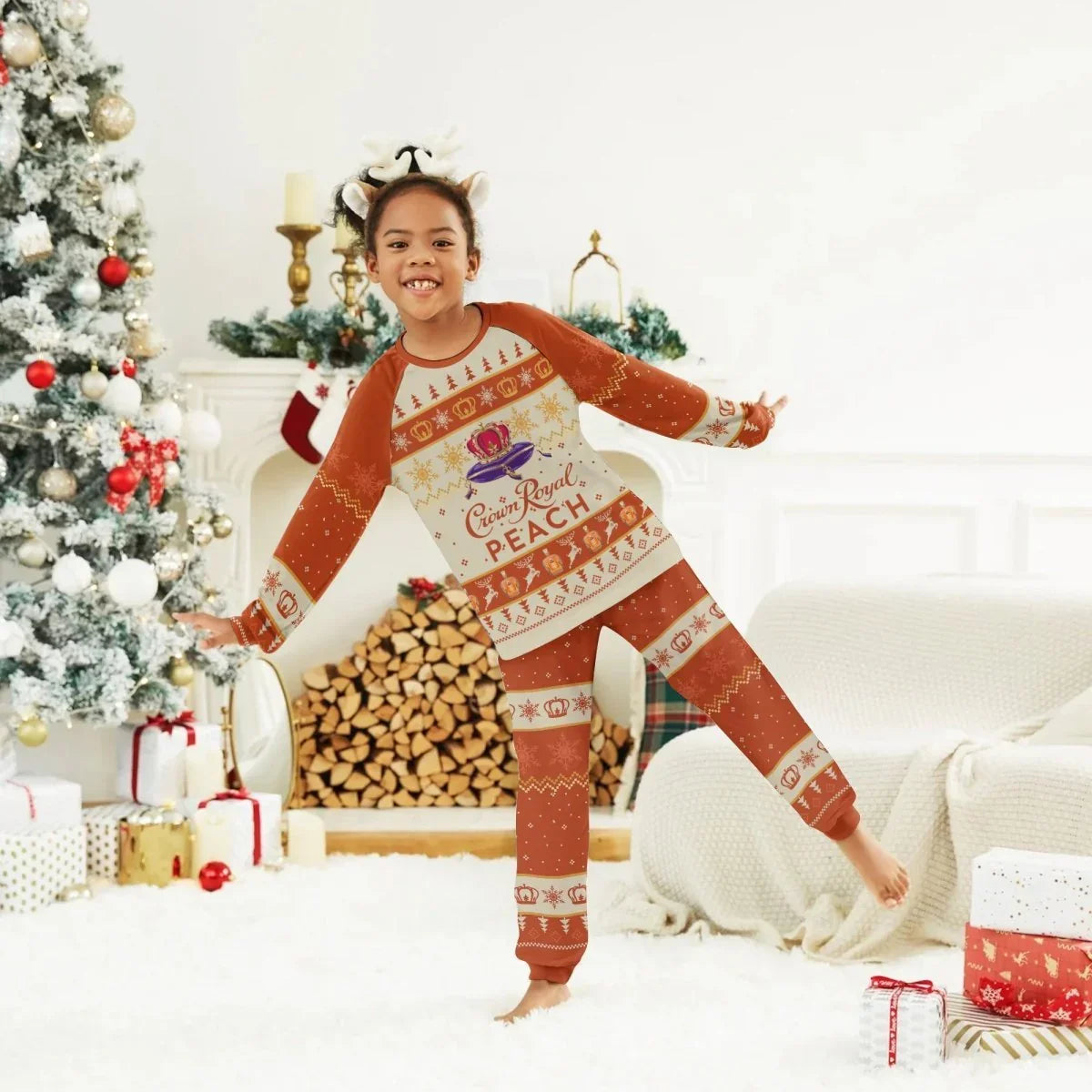 Child in Crown Royal Peach holiday pajamas and reindeer headband posing beside decorated Christmas tree and fireplace