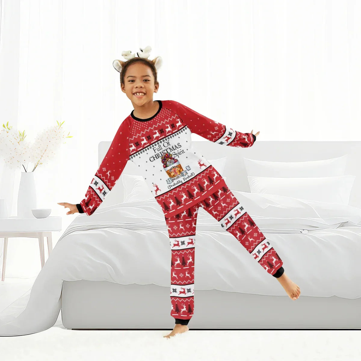 Smiling child in red and white Christmas pajamas with reindeer and holiday patterns standing on bed in bright room