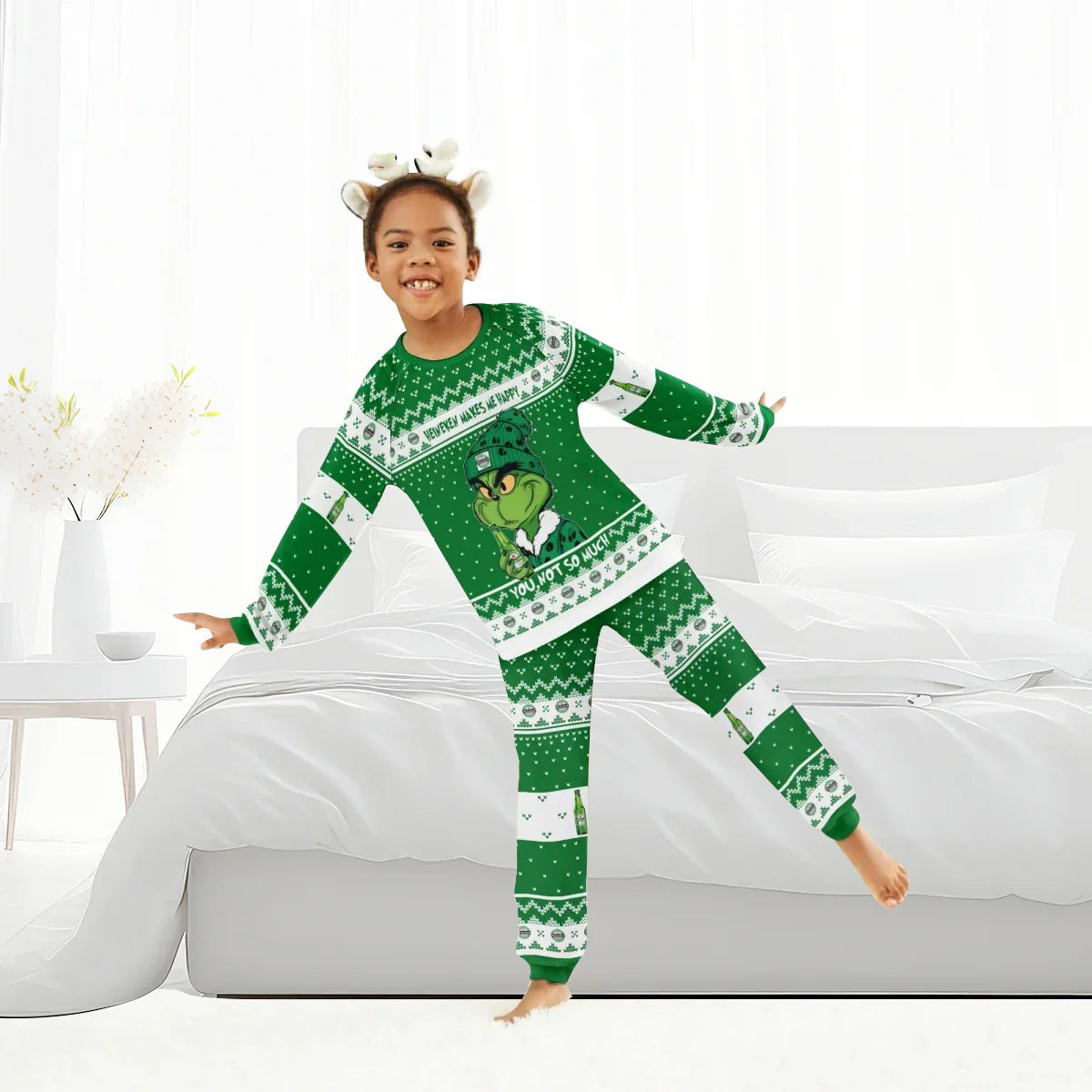 Smiling child in green Grinch-themed Christmas pajamas posing playfully in bright white bedroom