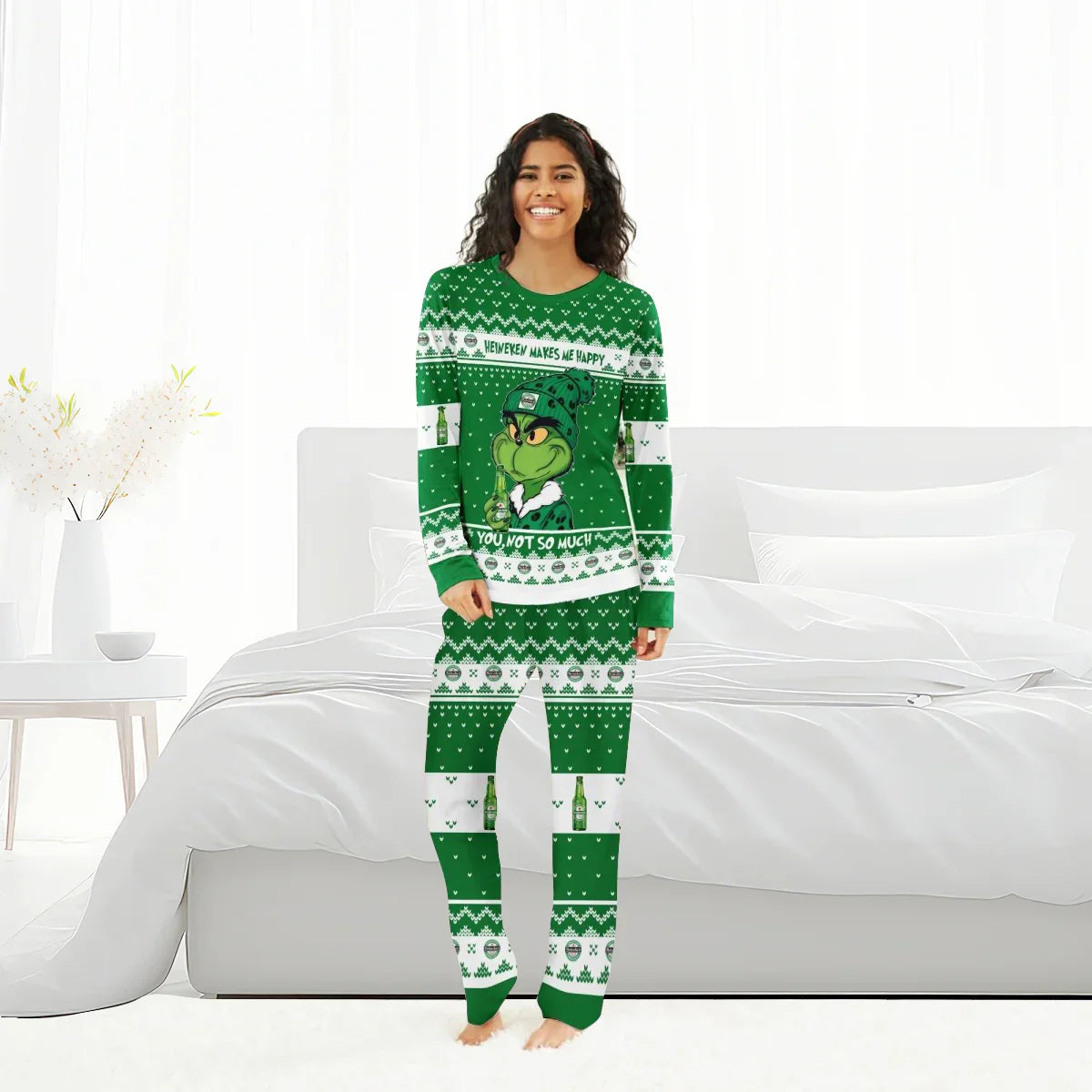 Smiling woman wearing green Heineken and Grinch-themed Christmas pajamas in bright white bedroom