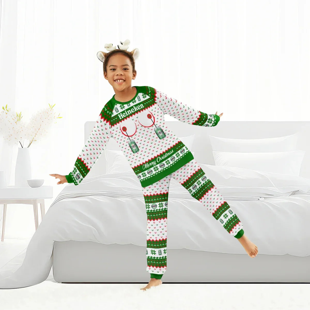 Child wearing Heineken Merry Christmas family pajamas with antler headband, posing by white bed