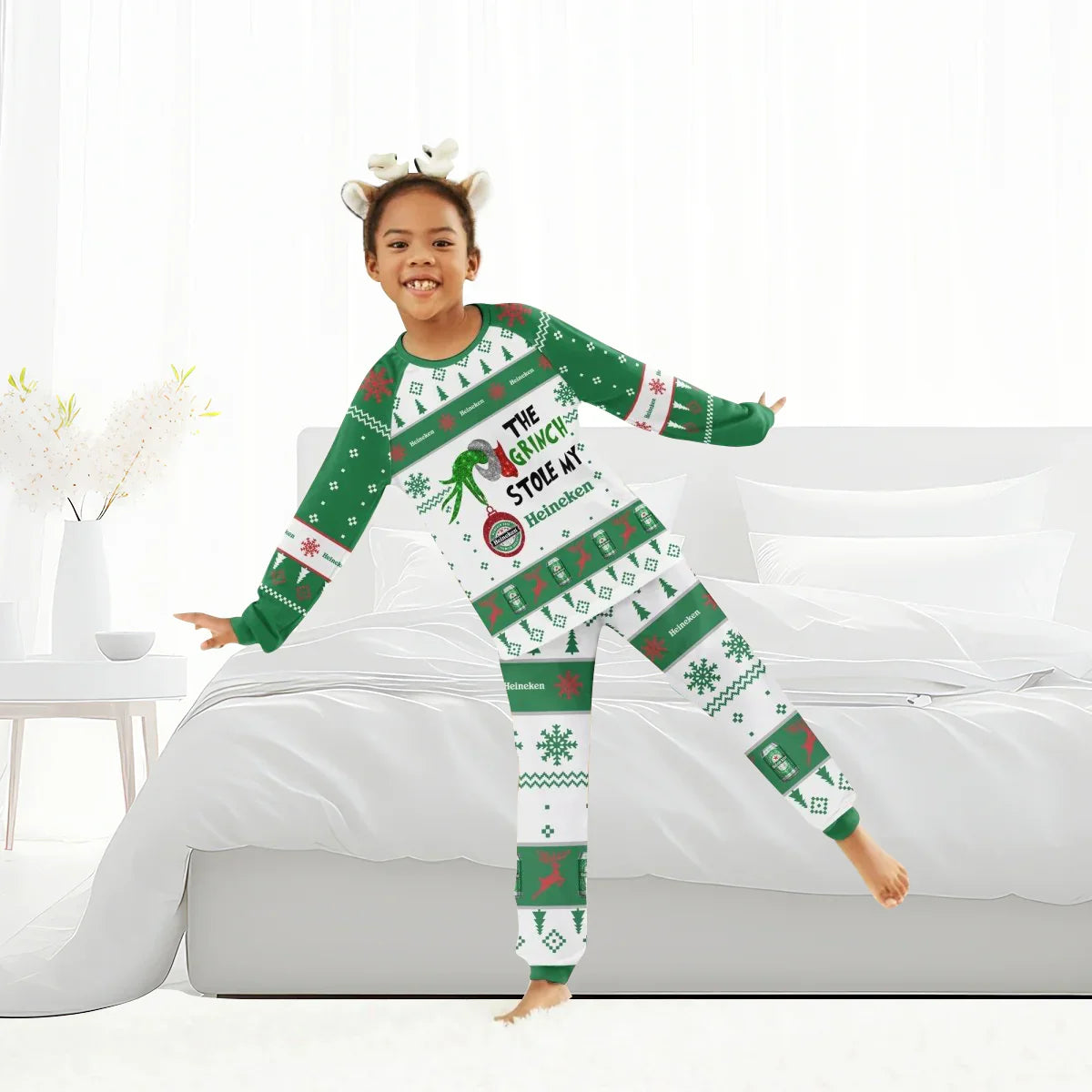 Child wearing green and white Heineken Grinch Christmas pajamas standing by a white bed in a bright room