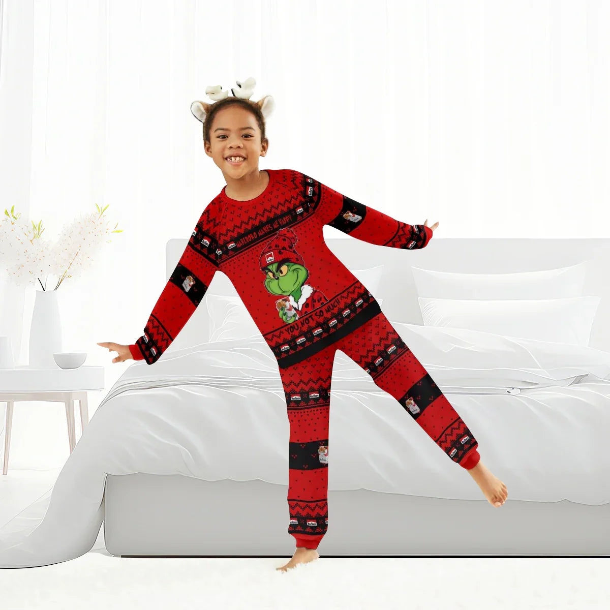 Child wearing red Grinch-themed Christmas pajamas posing in bright bedroom with white bed and decor