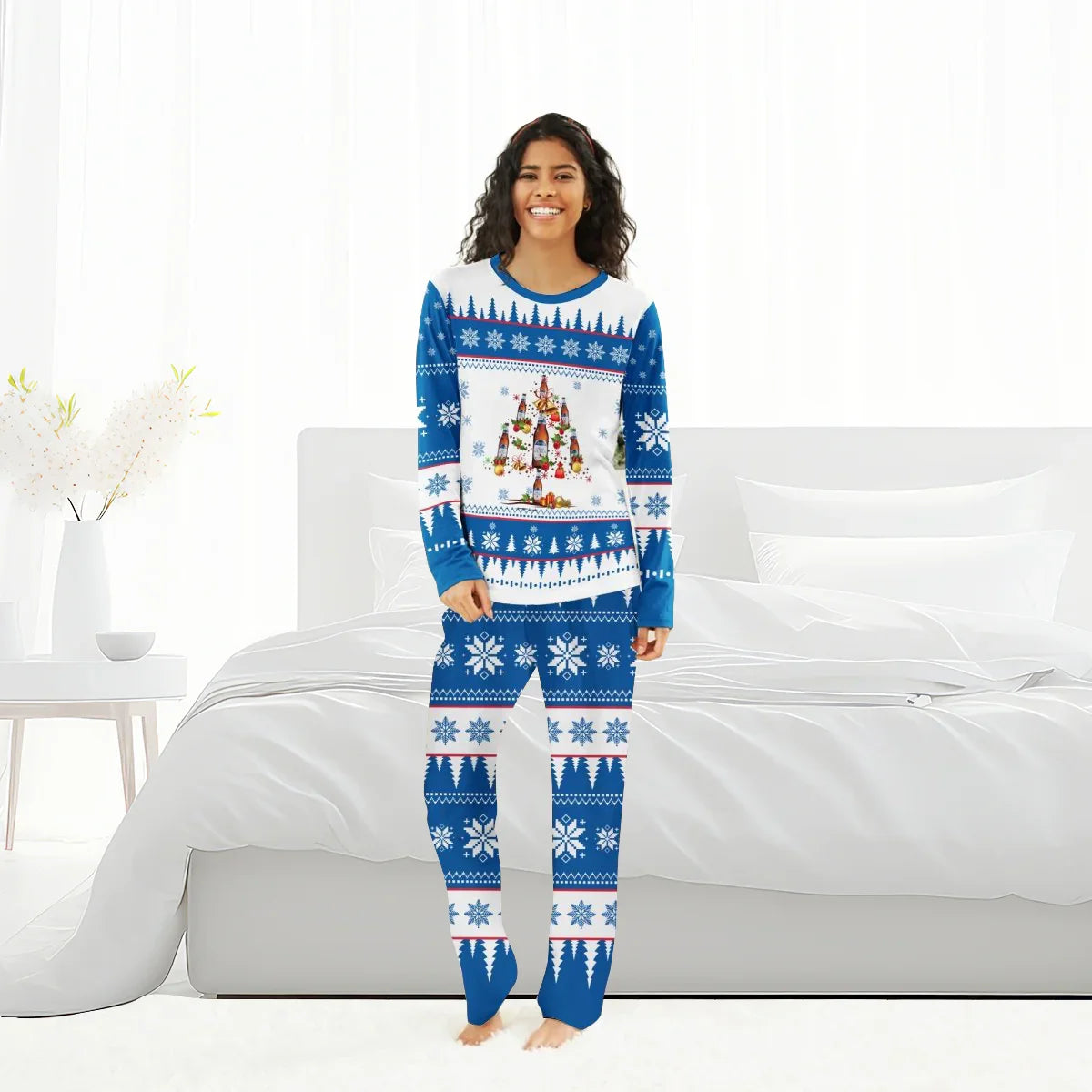 Smiling woman in blue and white Christmas tree and snowflake patterned holiday pajamas standing barefoot in modern white bedroom