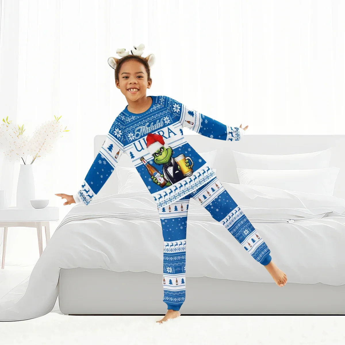 Child wearing blue Michelob Ultra Grinch Christmas holiday pajamas and reindeer antler headband on white bed