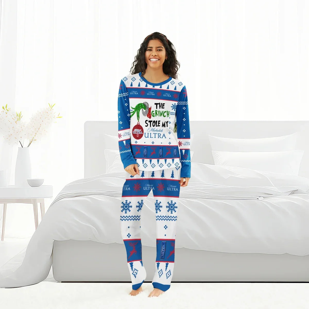 Smiling woman in blue and white Grinch-themed Christmas pajamas with Michelob Ultra branding, standing by a white bed