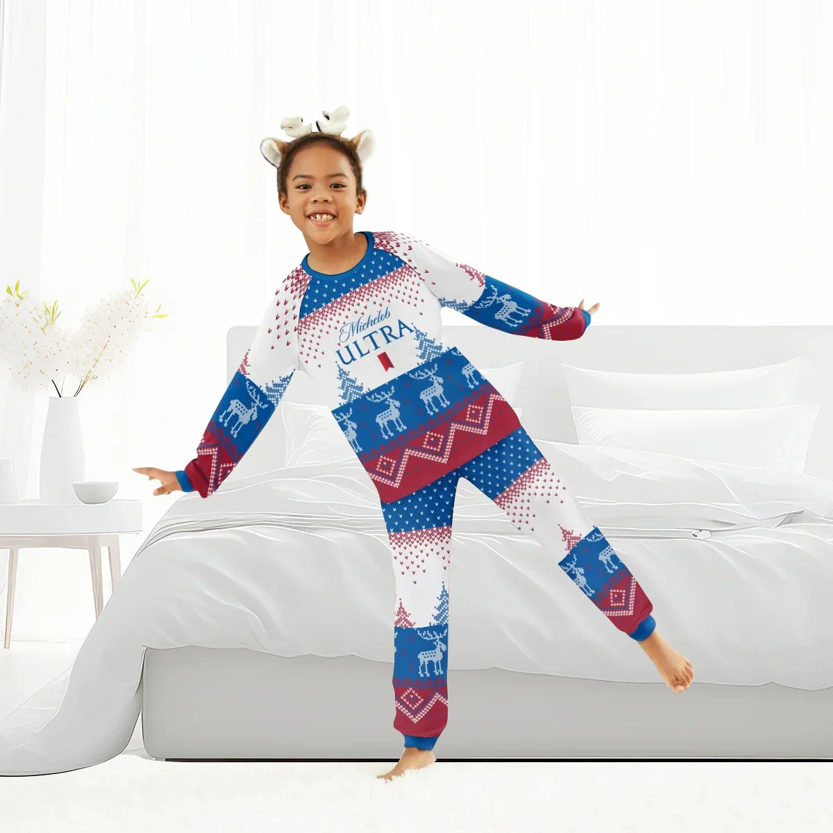 Smiling child in Michelob Ultra holiday reindeer pajamas and headband standing on bed in bright bedroom