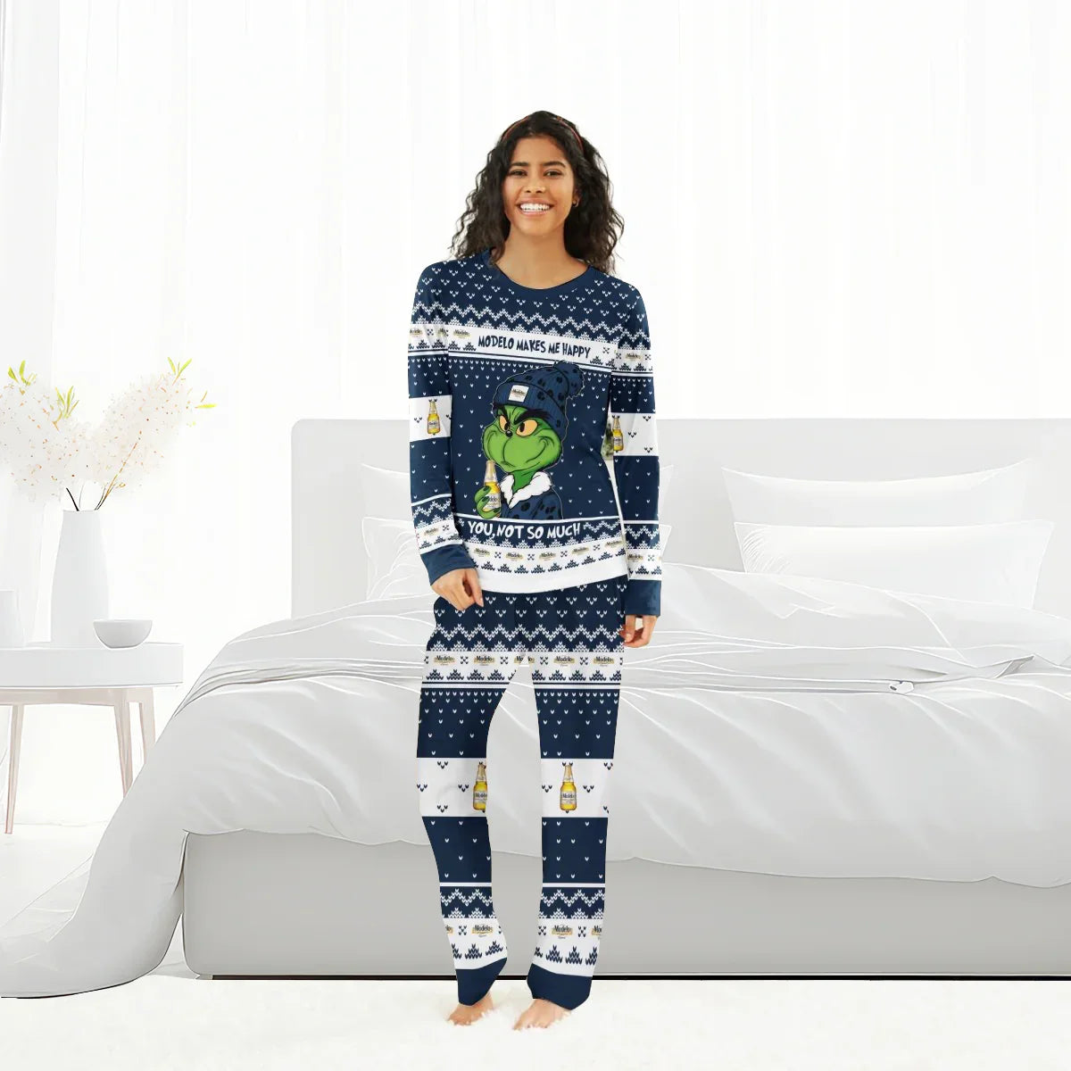 Smiling woman in navy blue Modelo and Grinch themed Christmas pajamas standing barefoot in bright bedroom