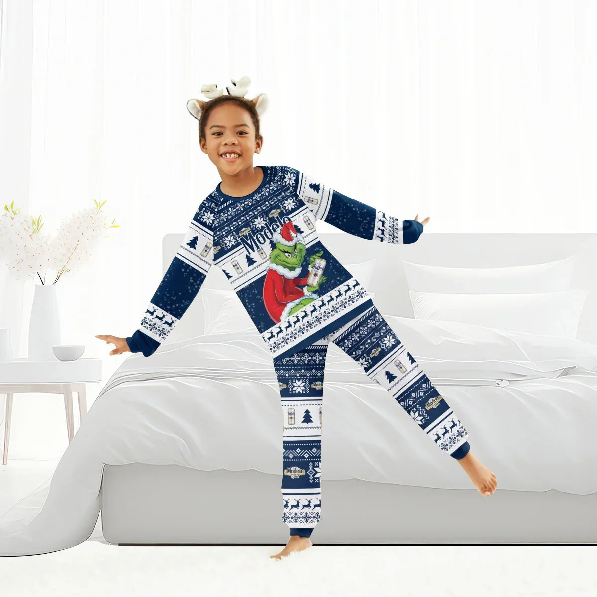 Child in festive blue Modelo Grinch Christmas pajamas playing on bed in bright bedroom