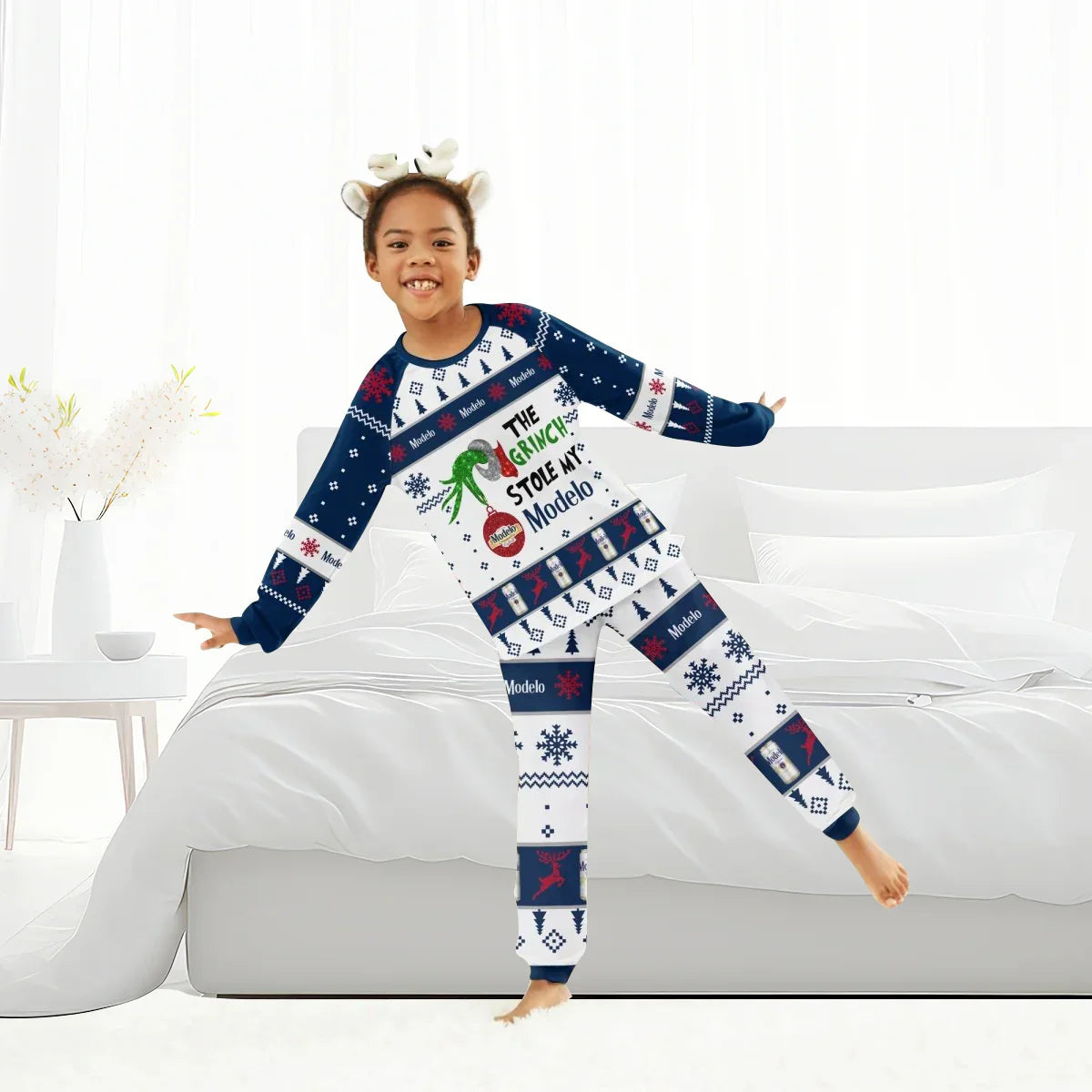 Child in festive Modelo and Grinch-themed holiday pajamas with antler headband in bright bedroom