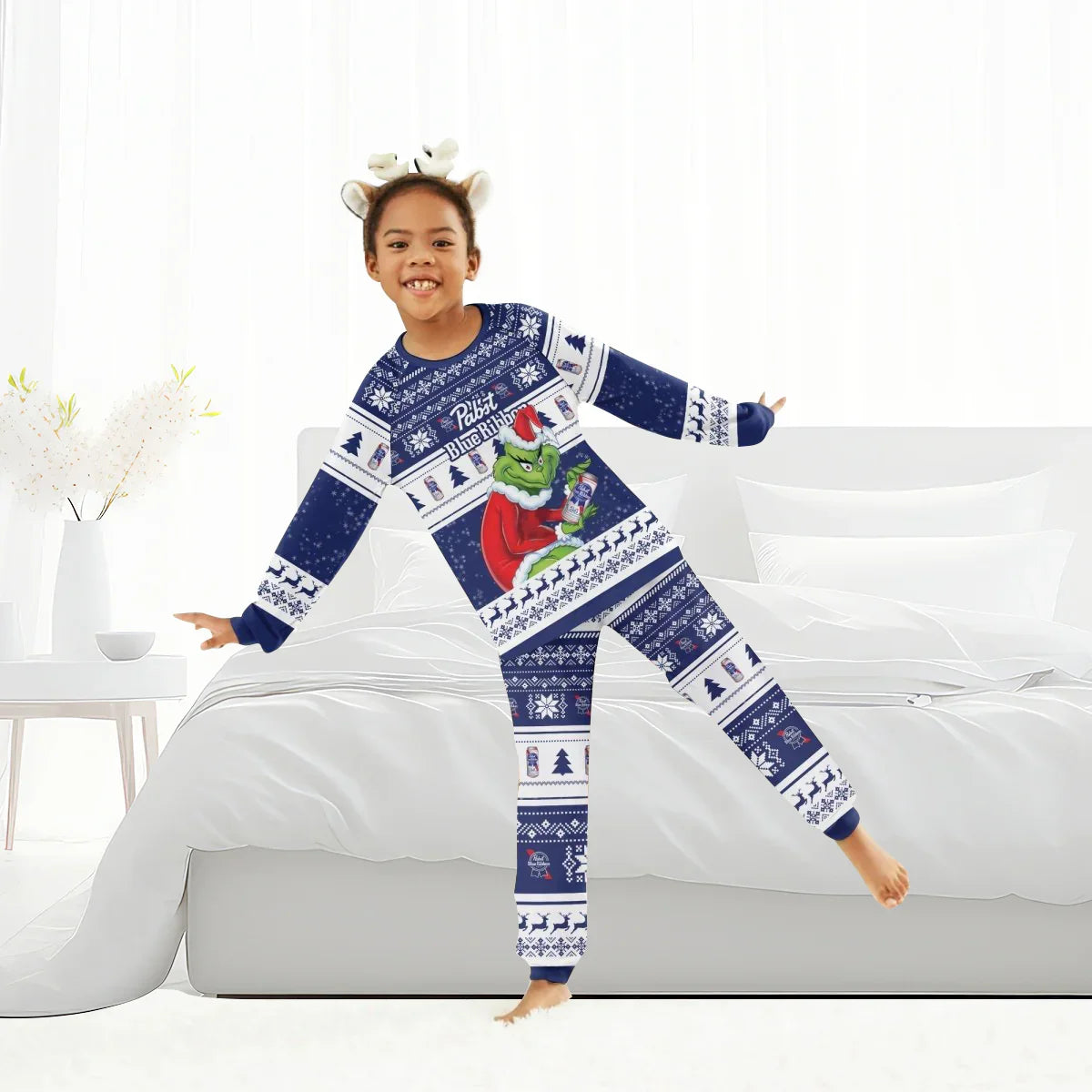 Child in navy blue Pabst Blue Ribbon Grinch Christmas pajamas and reindeer antler headband in bright bedroom
