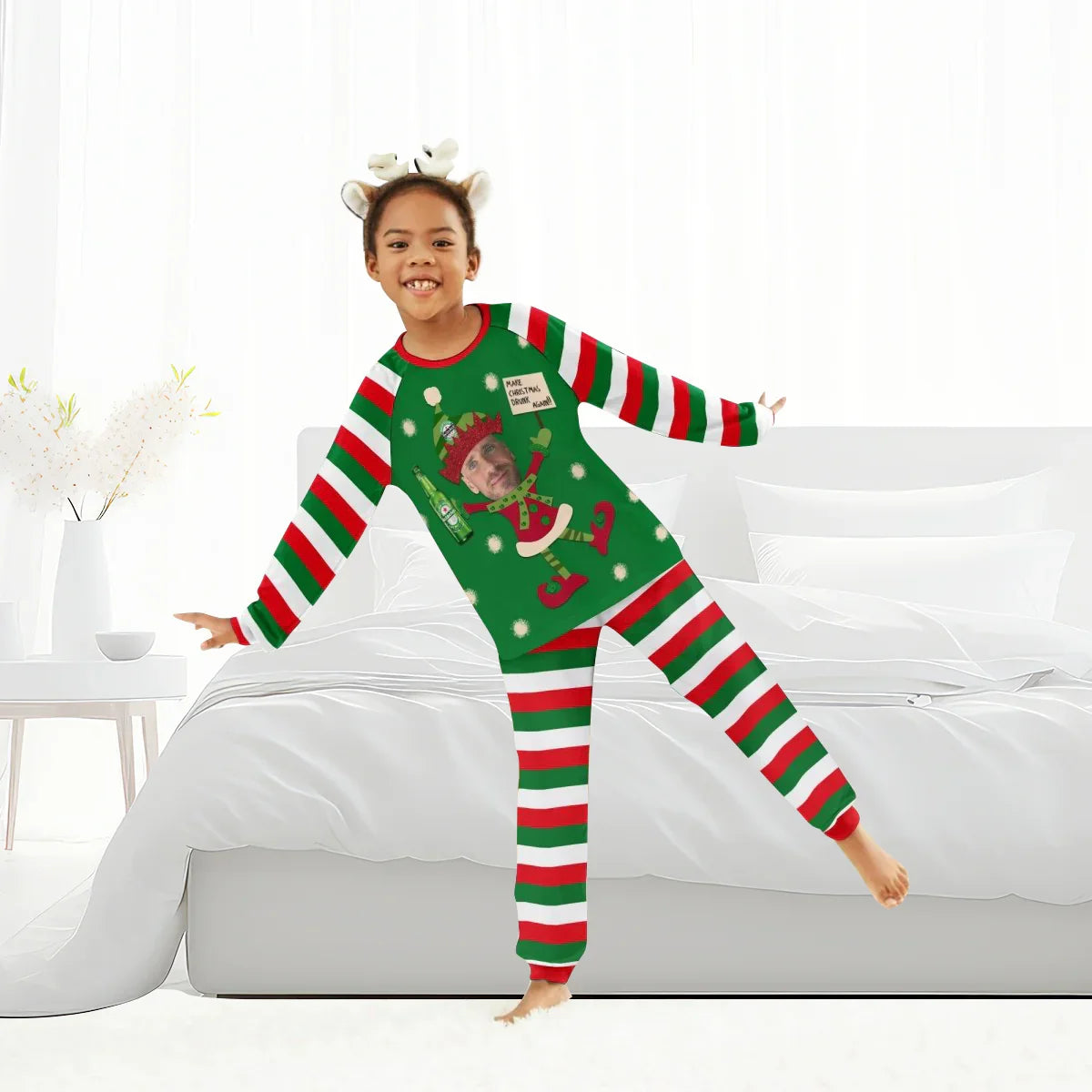 Child in green and red striped Christmas pajamas with elf print and antler headband in bright bedroom