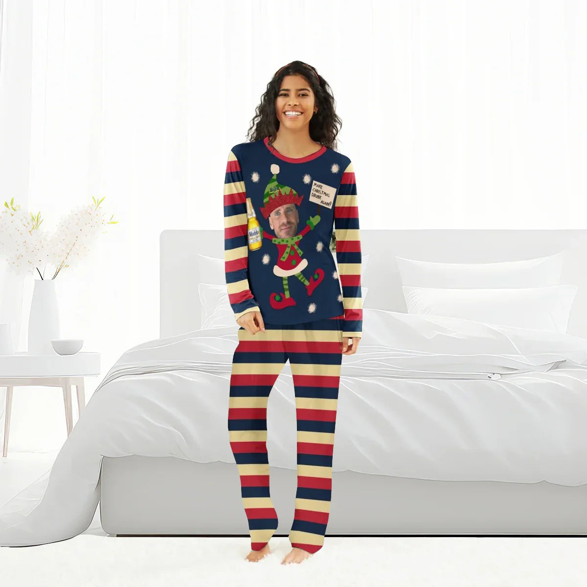 Smiling woman wearing striped Christmas pajamas with custom elf graphic holding Modelo beer in bedroom