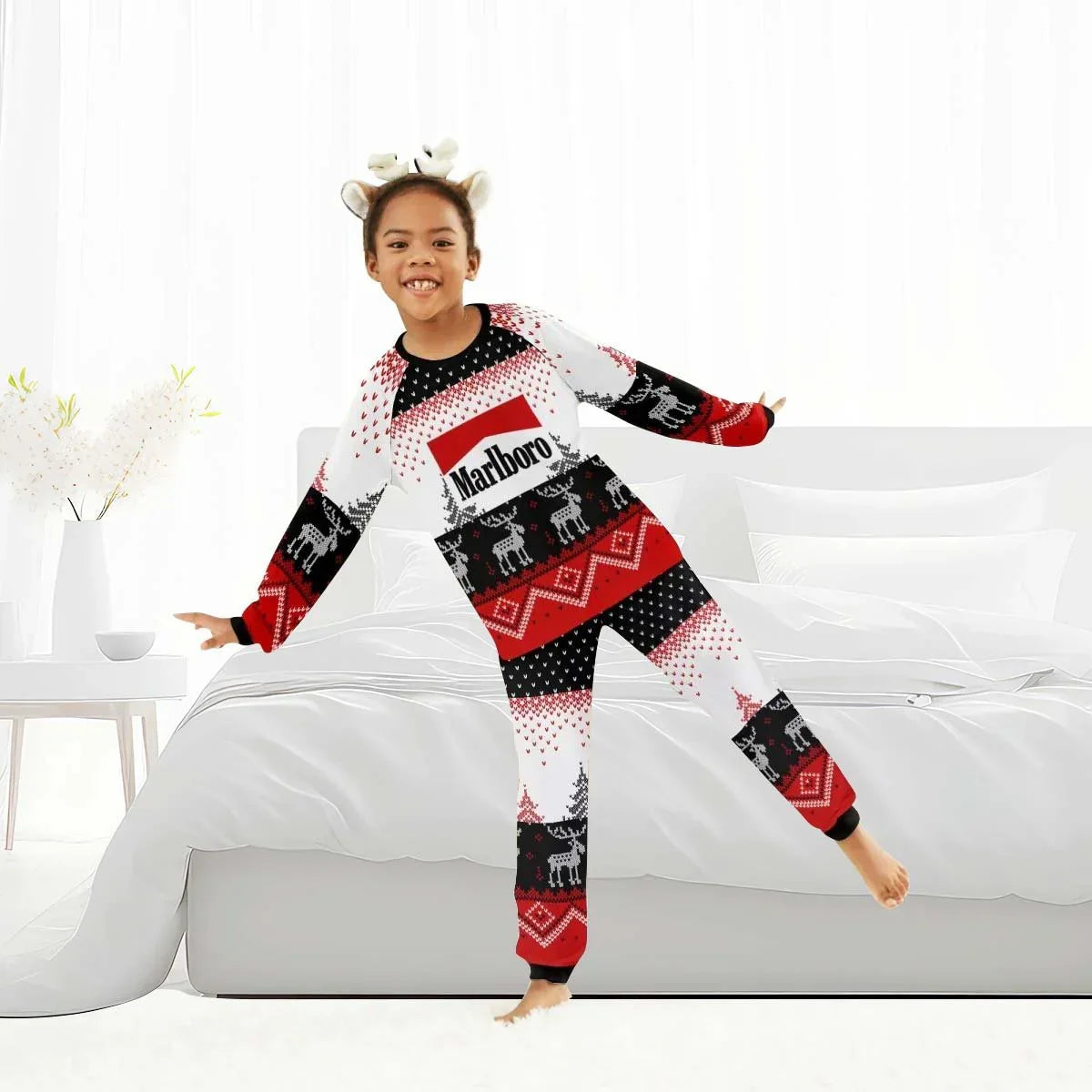 Child in red, black, and white Marlboro reindeer family winter pajamas jumping on a white bed in a bright room