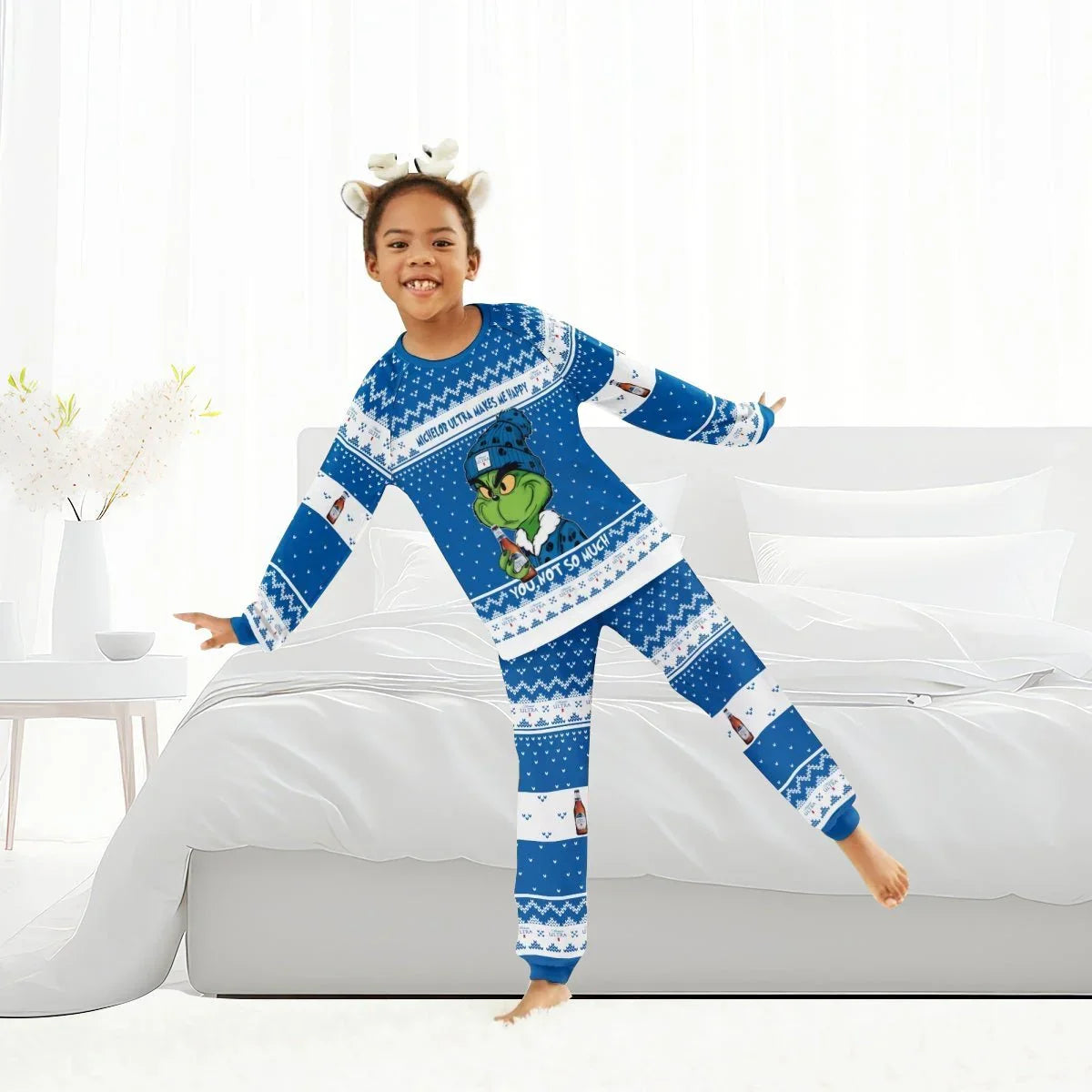 Child wearing blue Grinch-themed family pajamas with Michelob Ultra branding standing on white bed